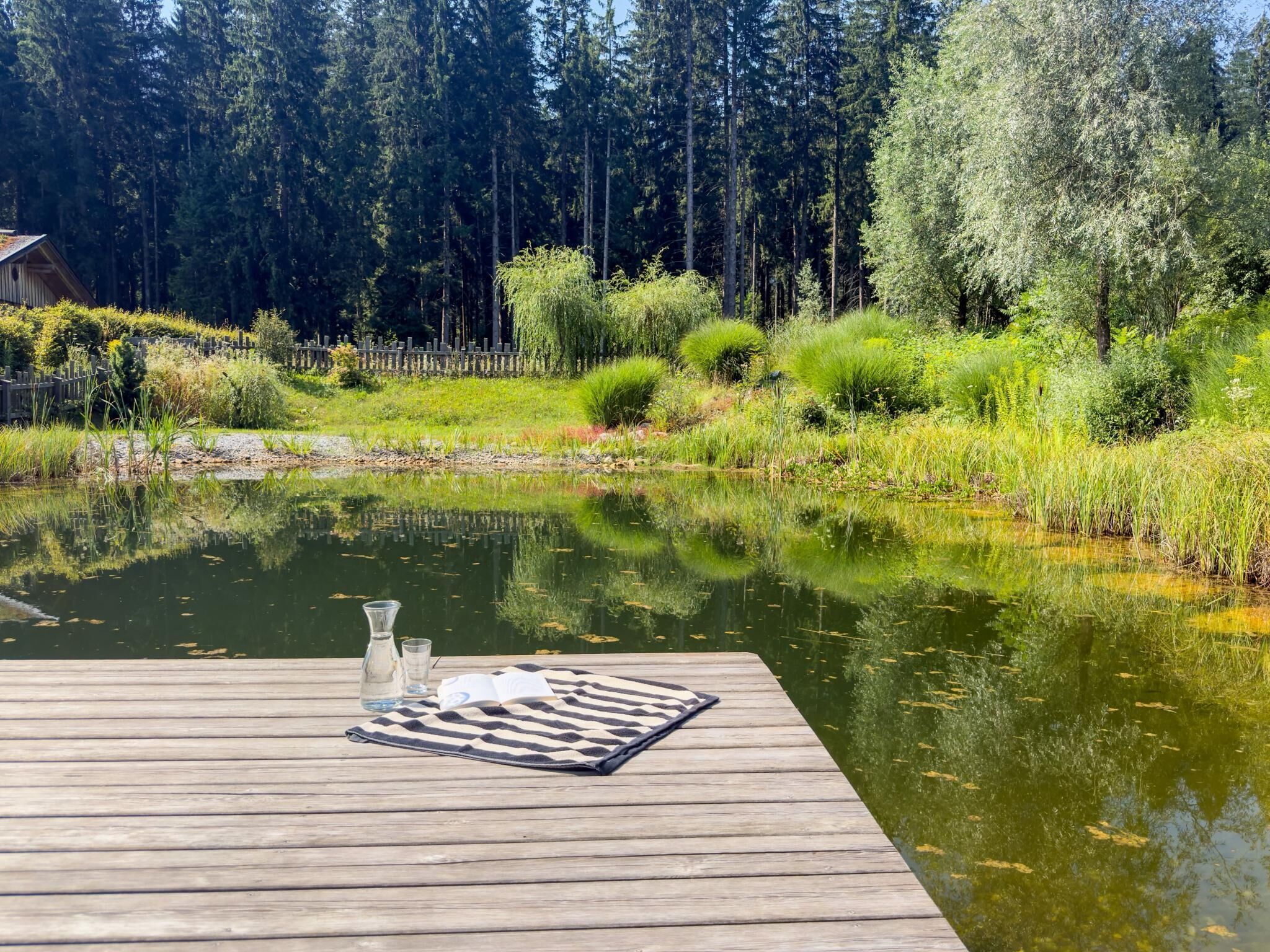 A wooden dock extending into a calm pond with a wooden fence and dense trees in the background.