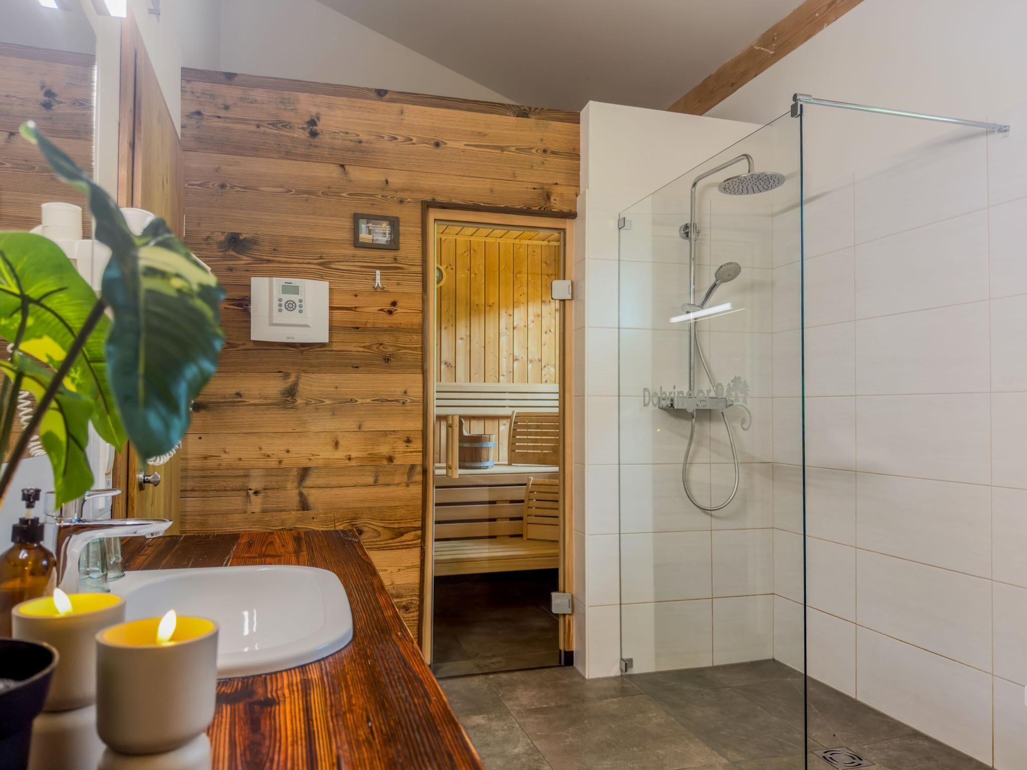 Ein Badezimmer mit Holzwand, einer Duschkabine aus Glas, einer hölzernen Sitzbank und einem weißen Waschbecken.