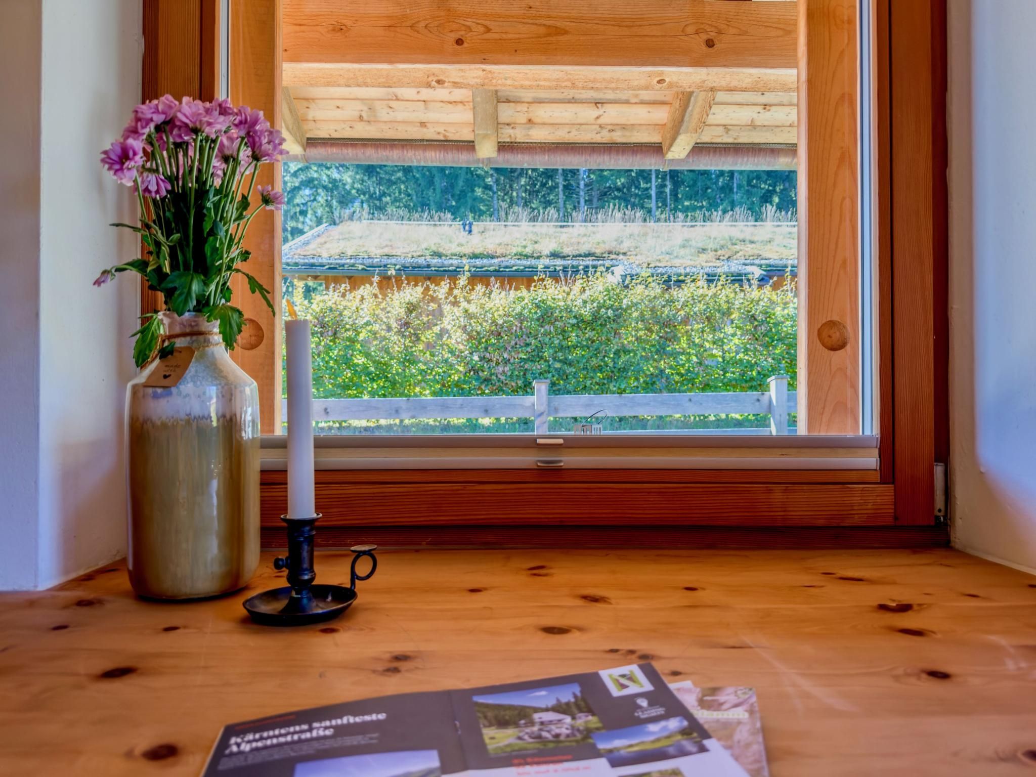 A wooden window sill with a vase of flowers and a candle, a brochure, and a view of a garden through the window.