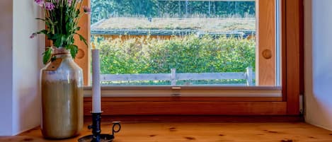 A wooden window sill with a vase of flowers and a candle, a brochure, and a view of a garden through the window.