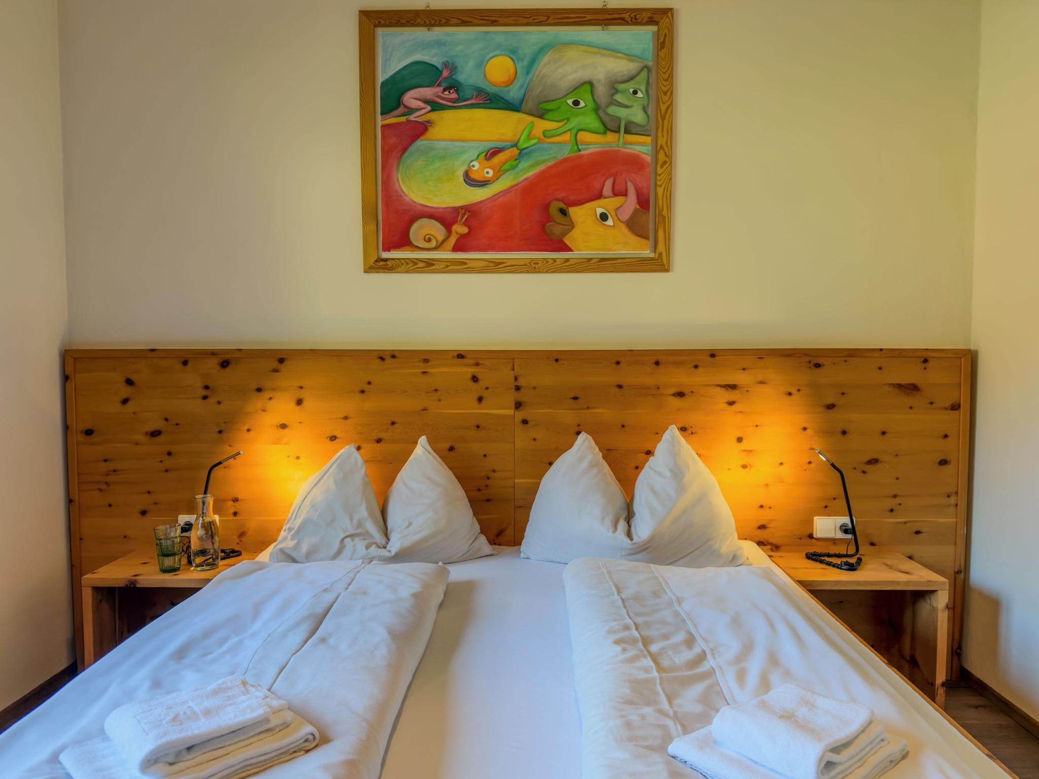 Un lit double avec du linge de lit blanc, deux tables de chevet, une tête de lit en bois et un tableau coloré au-dessus.