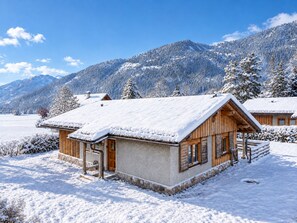 A wooden cabin with a snow-covered roof, surrounded by a snowy landscape and mountains.