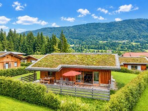 A modern wooden cabin with a green roof, a deck with outdoor furniture, and a scenic mountain view.