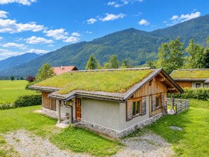 A traditional wooden house with a grass roof, set in a lush green valley with mountains in the background.