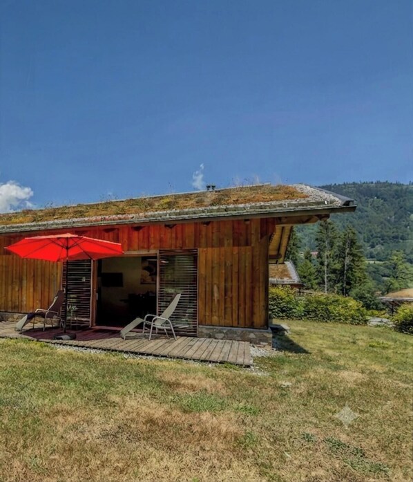 A wooden cabin with a red umbrella, two chairs, and a view of a forested mountain.