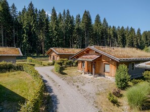 Una cabaña de madera con camino de grava, rodeada de un bosque frondoso y un cielo despejado.