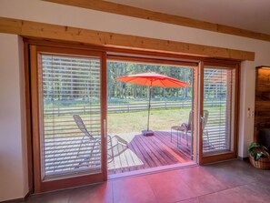 A sliding glass door leading to an outdoor deck with a red umbrella, metal chairs, and a view of a forested area.
