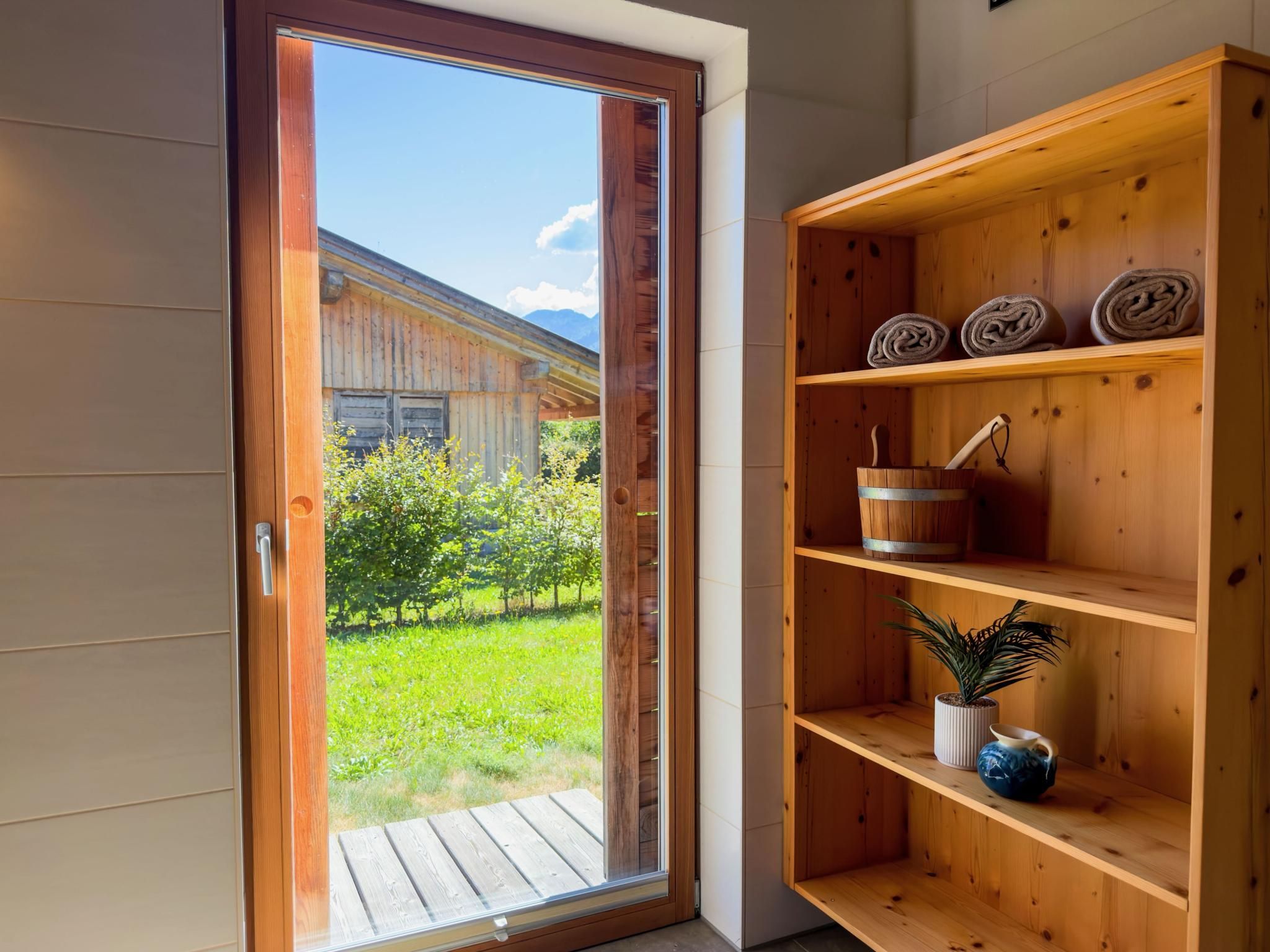 A wooden shelf with rolled towels and a bucket, a potted plant, and a wooden door leading to a garden.