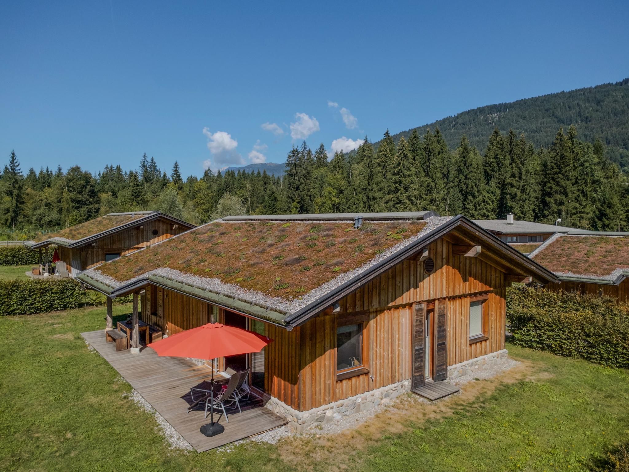 Une cabane en bois avec un toit en pente, une terrasse couverte avec un parasol rouge et une vue sur une chaîne de montagnes boisées.