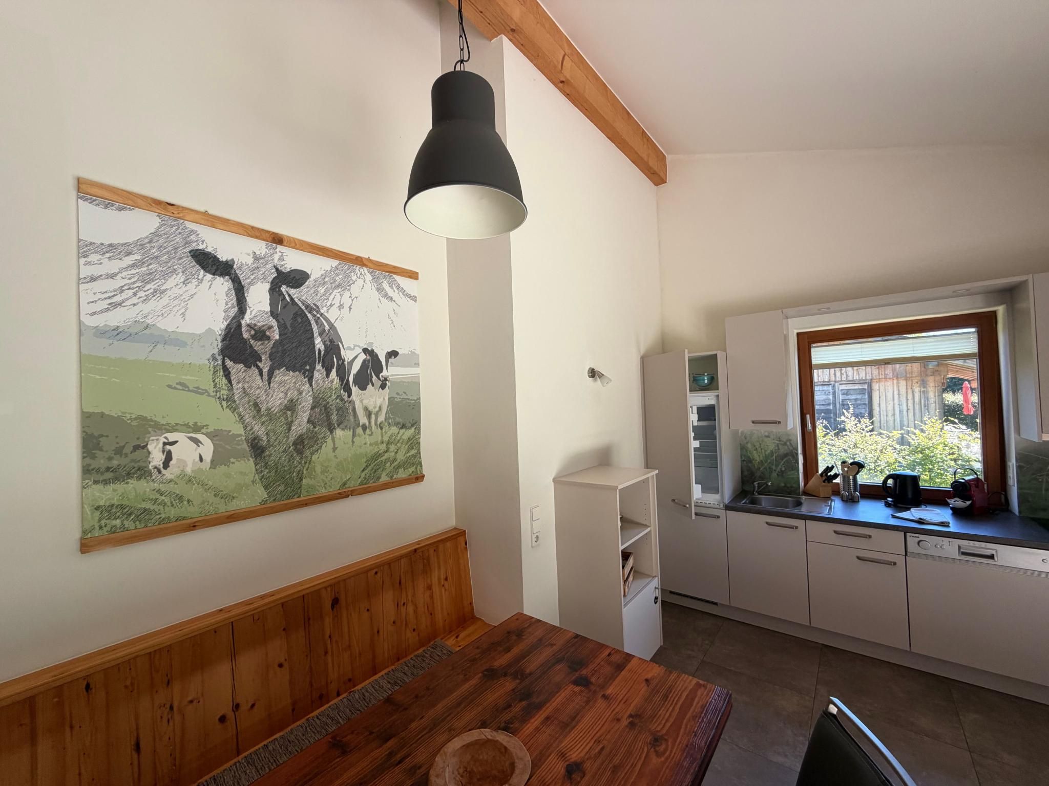 Een eetgedeelte met een houten tafel, een schilderij van koeien en een keuken met een raam.