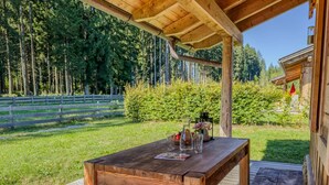 Een houten tafel met een glazen blad, omringd door een houten terras, met op de achtergrond een bos en een heldere lucht.