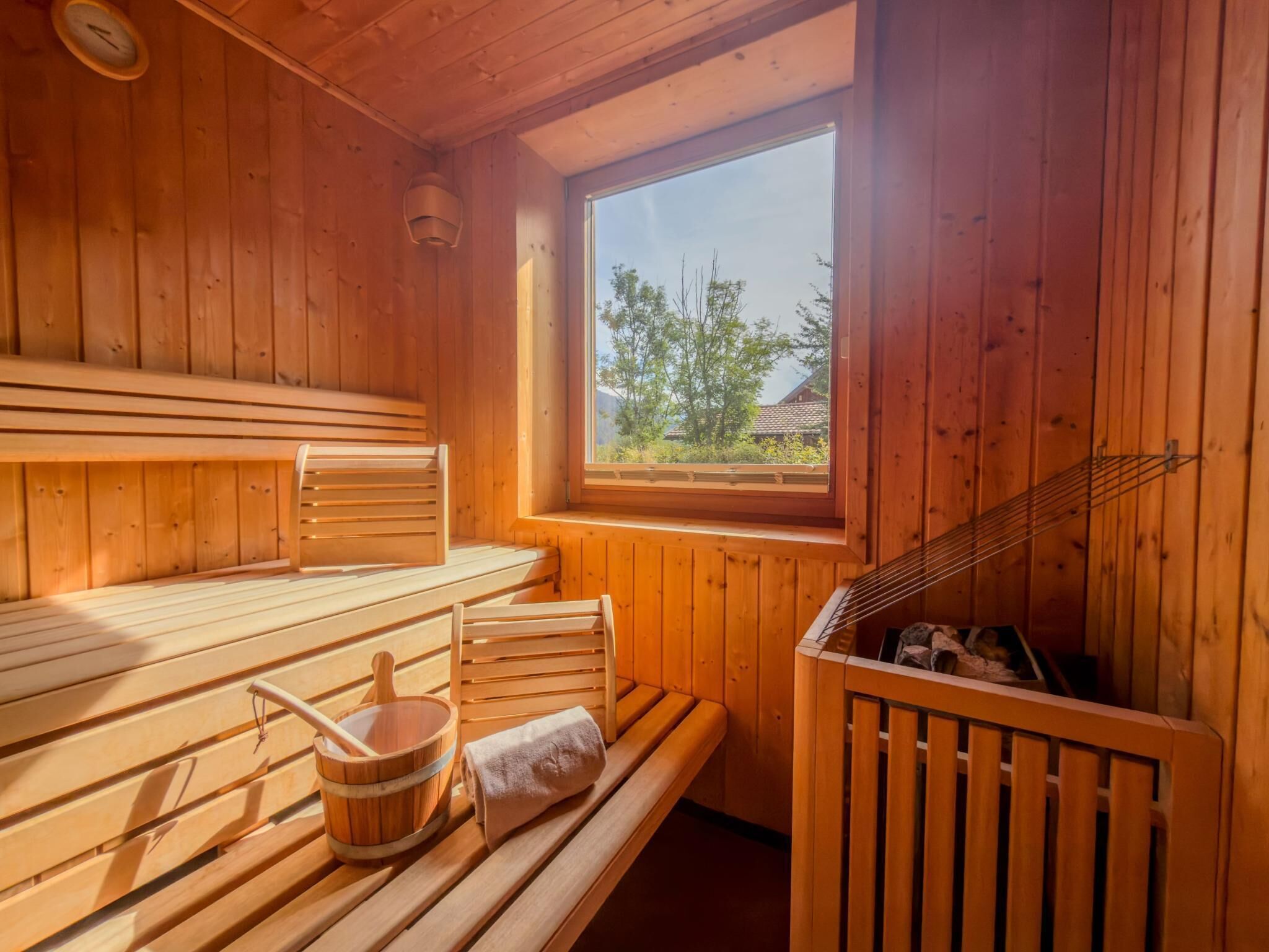 A sauna room with wooden benches, a bucket, and a window with a view of trees and a house.