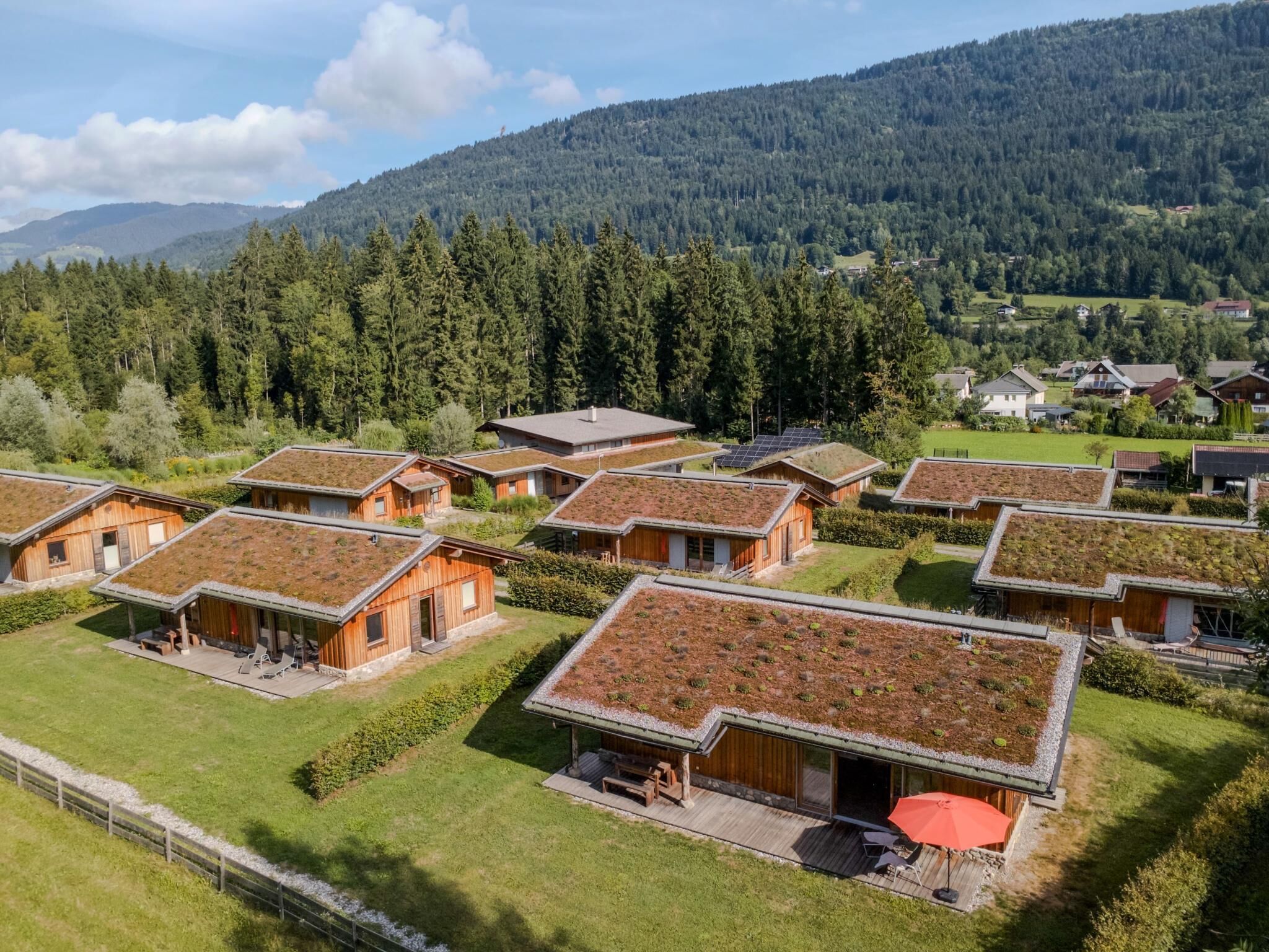 A cluster of wooden cabins with sloped roofs, surrounded by a forested landscape and hills in the background.