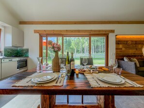 A dining area with a wooden table set for two, a kitchen with modern appliances, and a view of a forest through large windows. - Chalet with sauna near the Nassfeld region (Hermagor-Pressegger See)