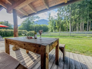 Eine Holzterrasse mit Esstisch, die auf eine grüne Fläche mit Bäumen und einen Berg im Hintergrund blickt.