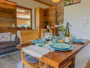 A wooden dining table set with plates, glasses, and a bottle, with a sofa and window in the background.