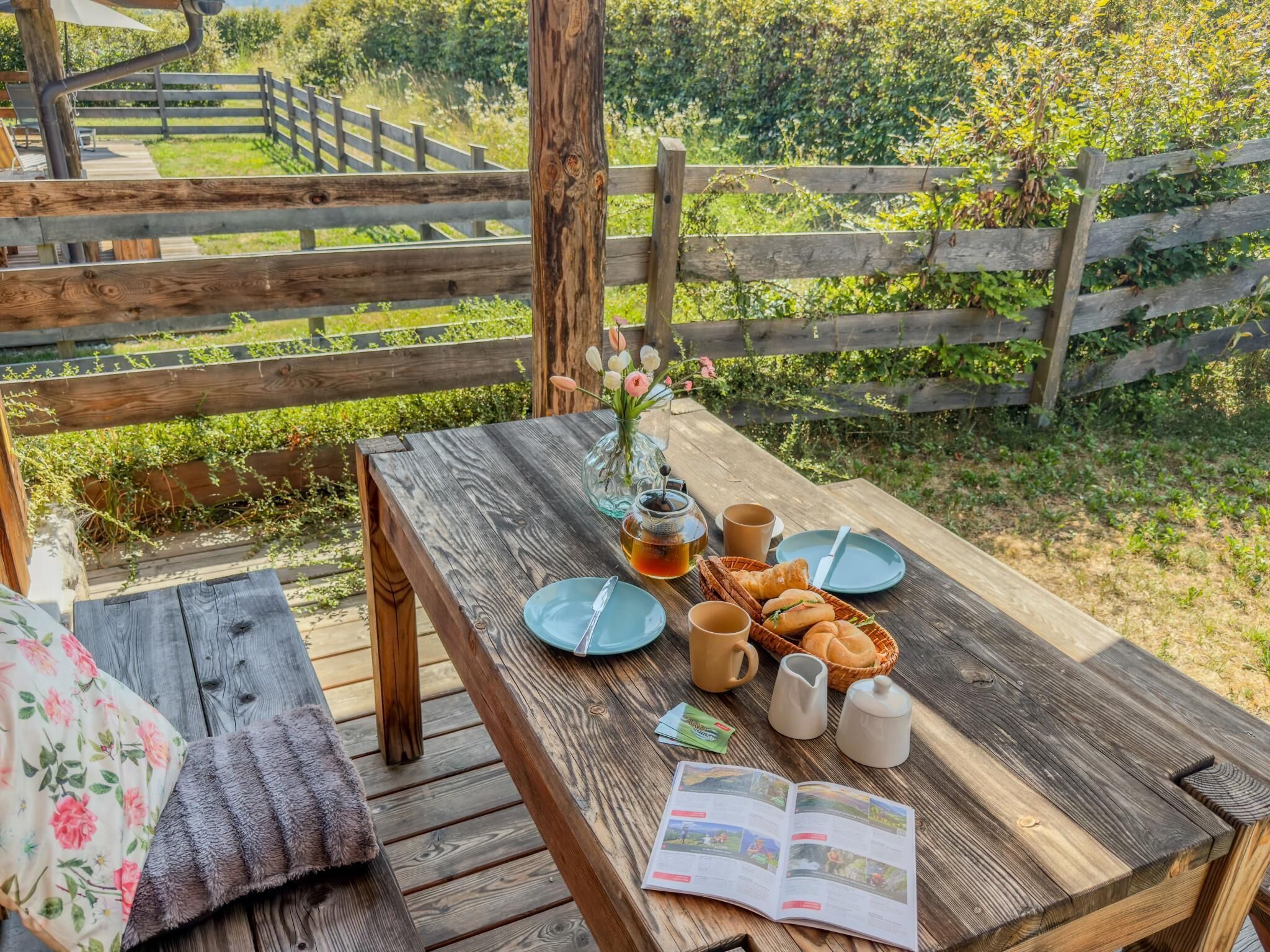 Une table de pique-nique en bois, installée à l’extérieur, avec un livre, une tasse, une théière et un panier de pain.