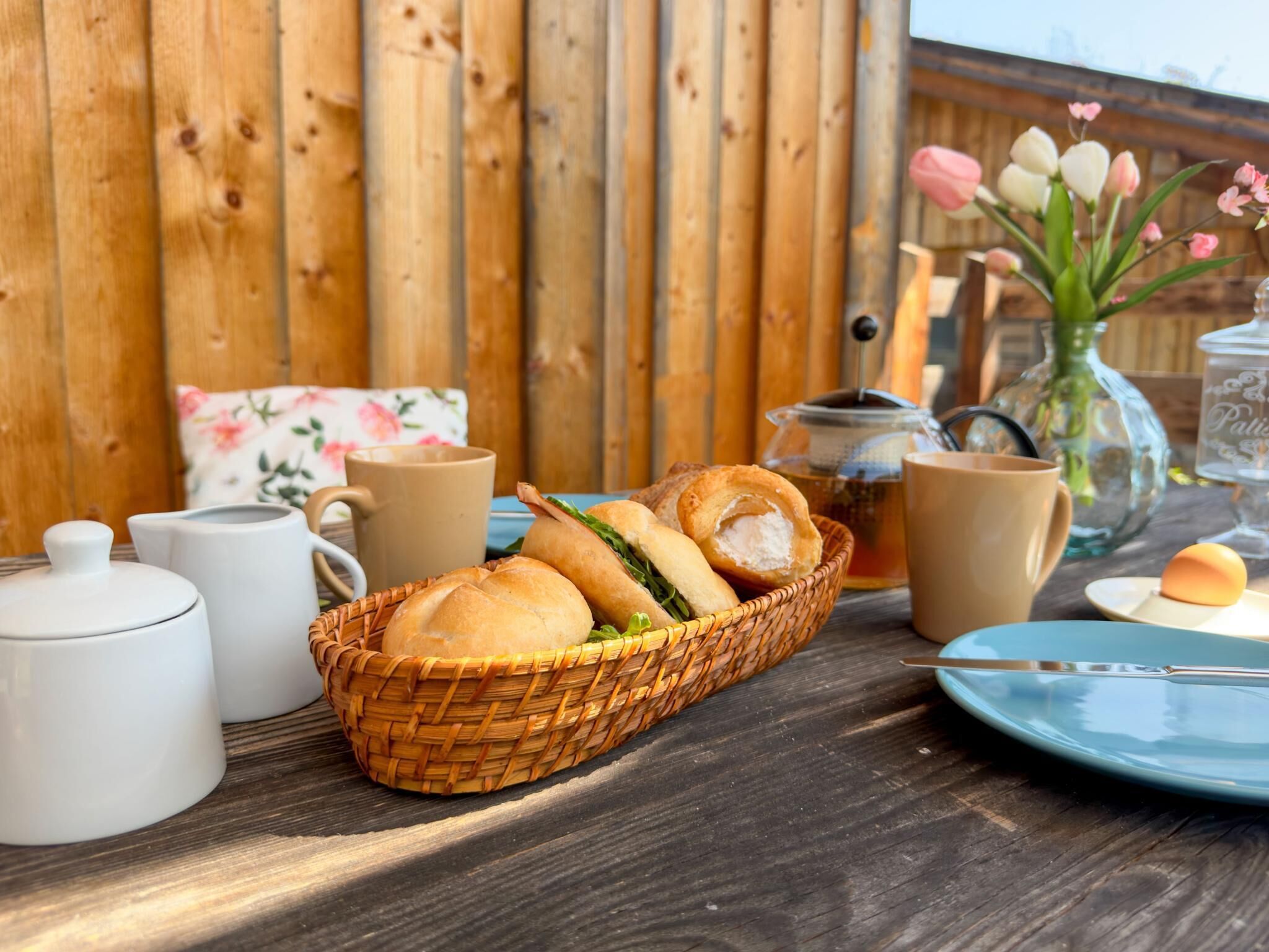 A wooden table with a basket of sandwiches, a teapot, and a cup.