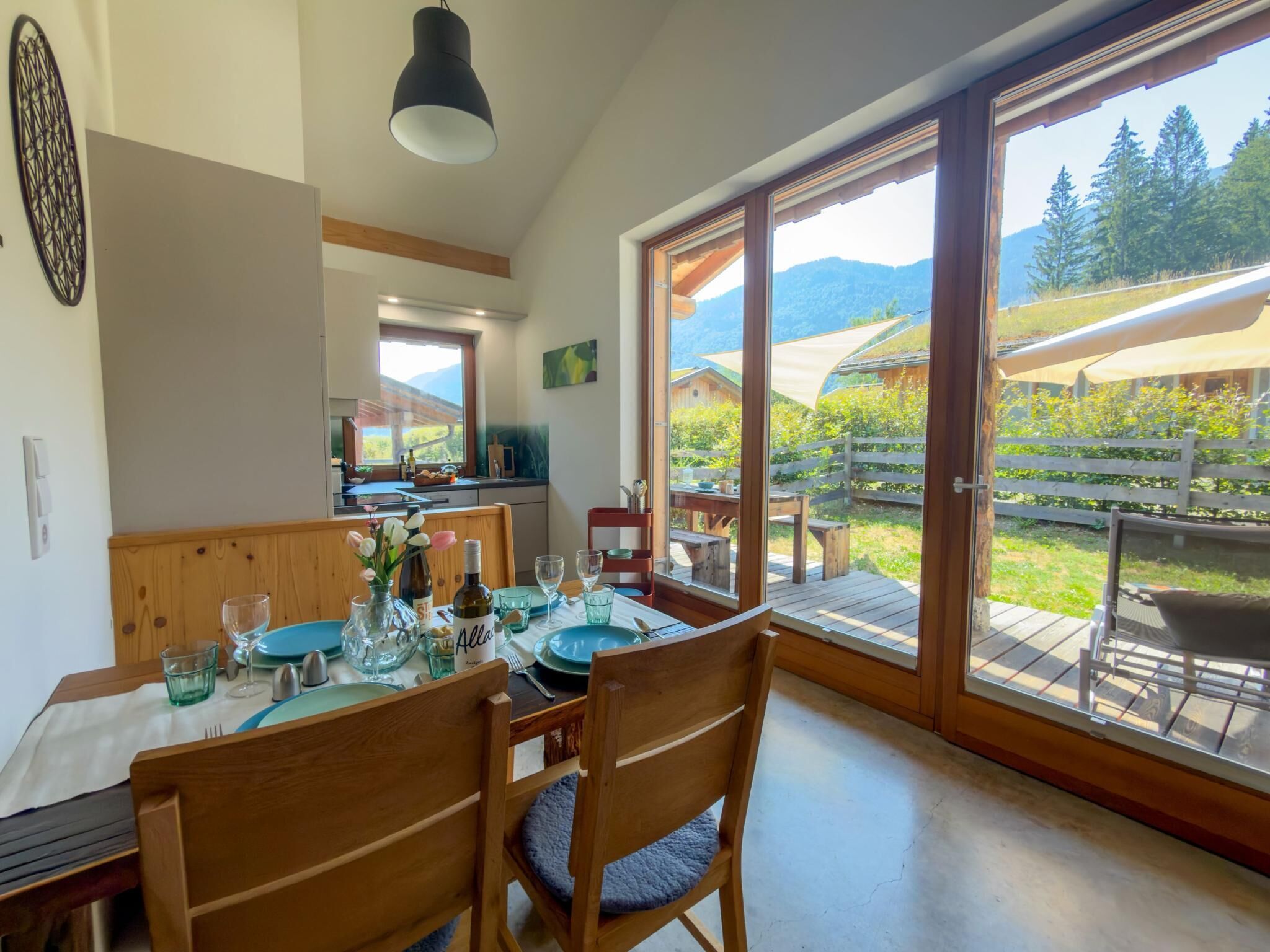 A dining area with a table set for two, wooden chairs, a large window with a view of the outdoors, and a hanging light fixture.