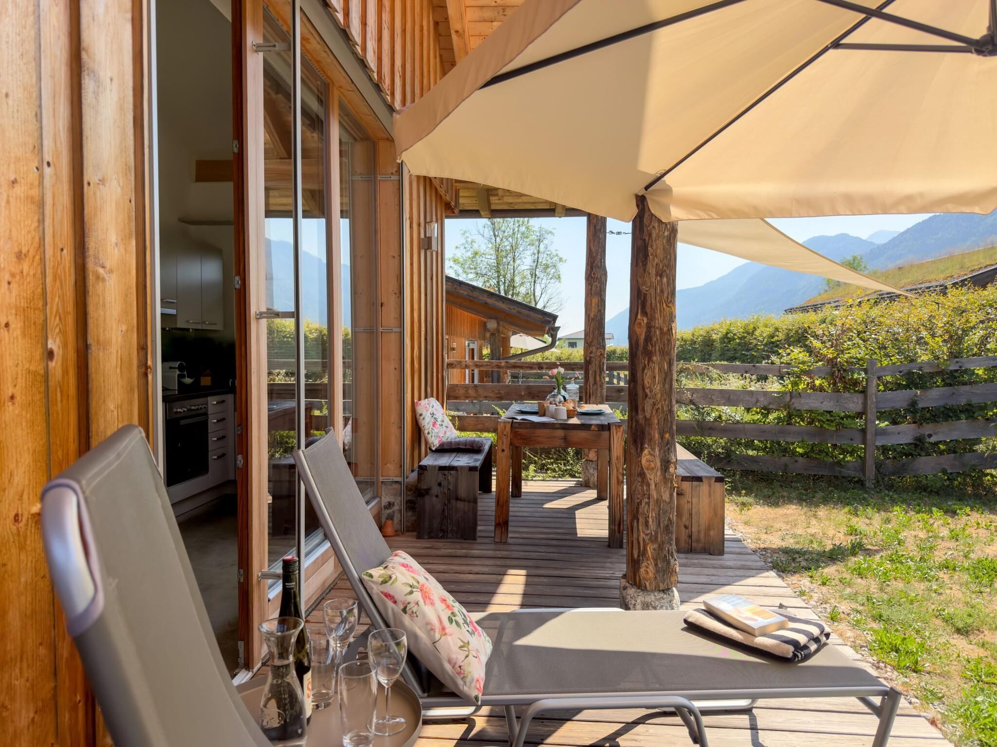 A wooden deck with a table, chairs, and a lounge chair, overlooking a vineyard and mountains.