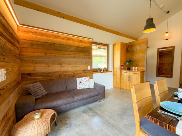 A cozy room with a sofa, a small table, a dining table with chairs, wooden walls, and a window.