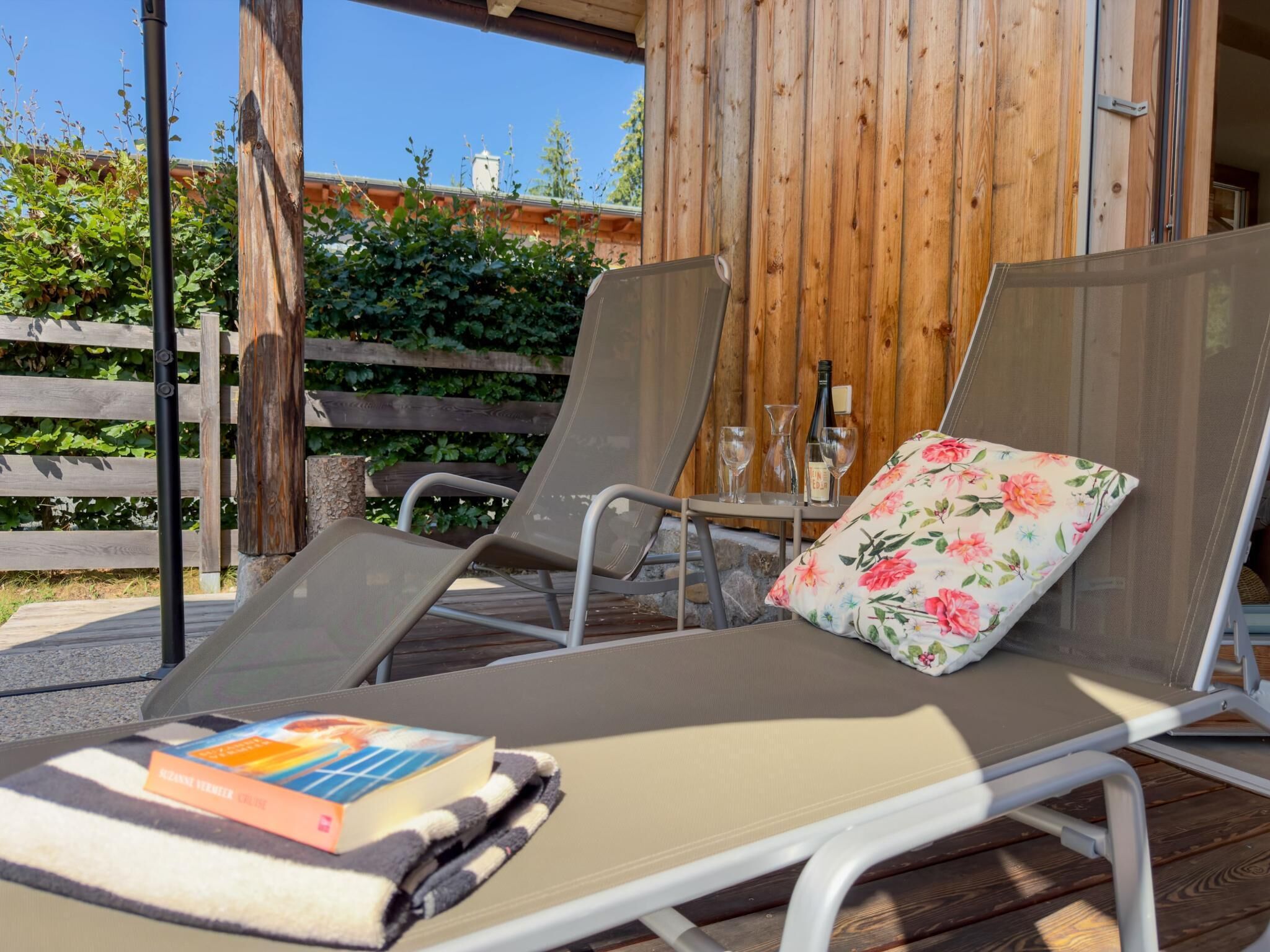 Een overdekt terras met twee comfortabele stoelen, een tafel met een fles en glazen, en een boek op een kussen.