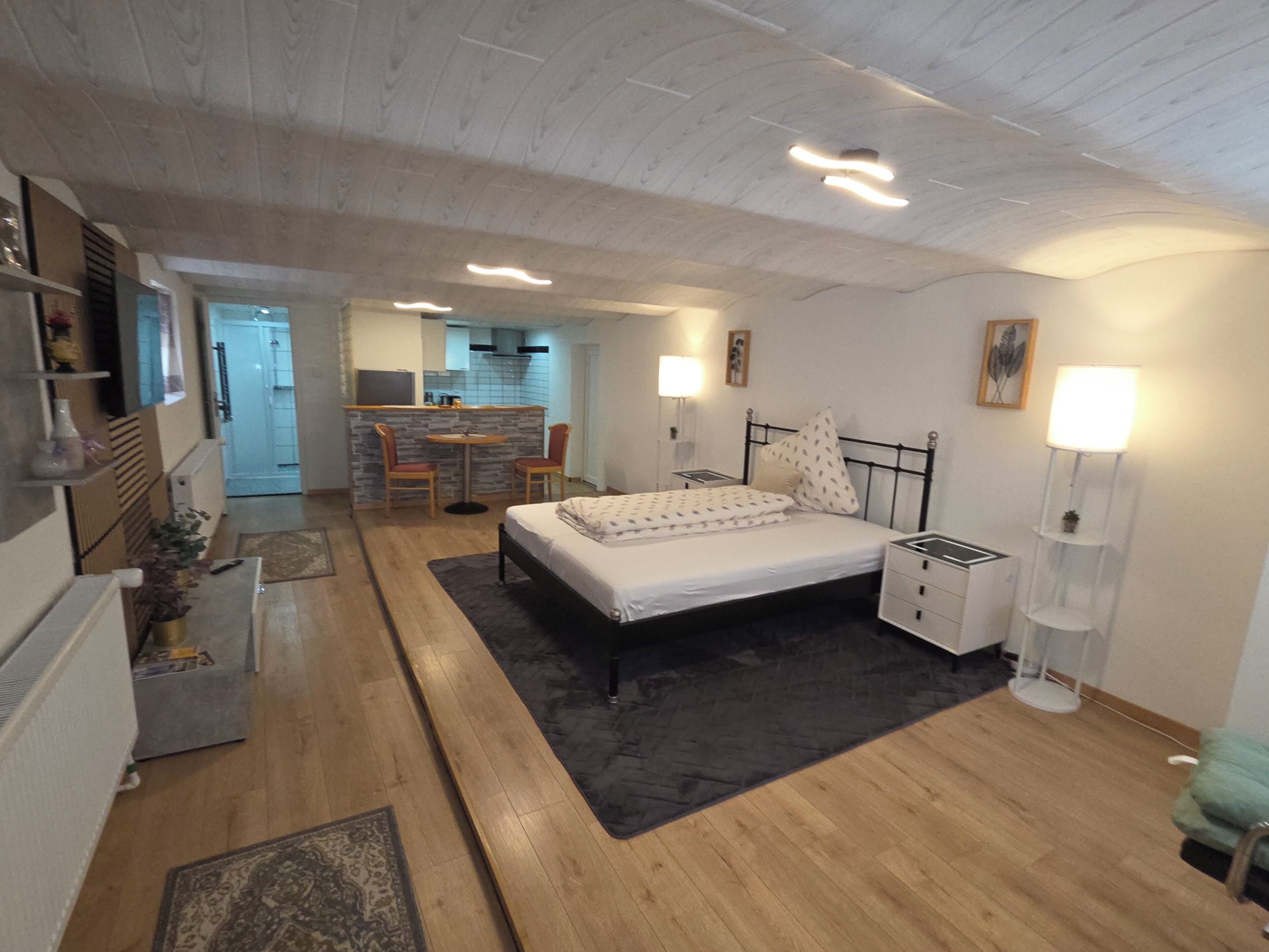 A bedroom with a bed, bedside tables, a desk, and a chair. There is a kitchenette in the background.