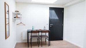 Ein kleiner Raum mit einem Tisch, zwei Stühlen, einer schwarzen Tür, einem weißen Heizkörper, einem Kleiderhaken und einem Spiegel.
