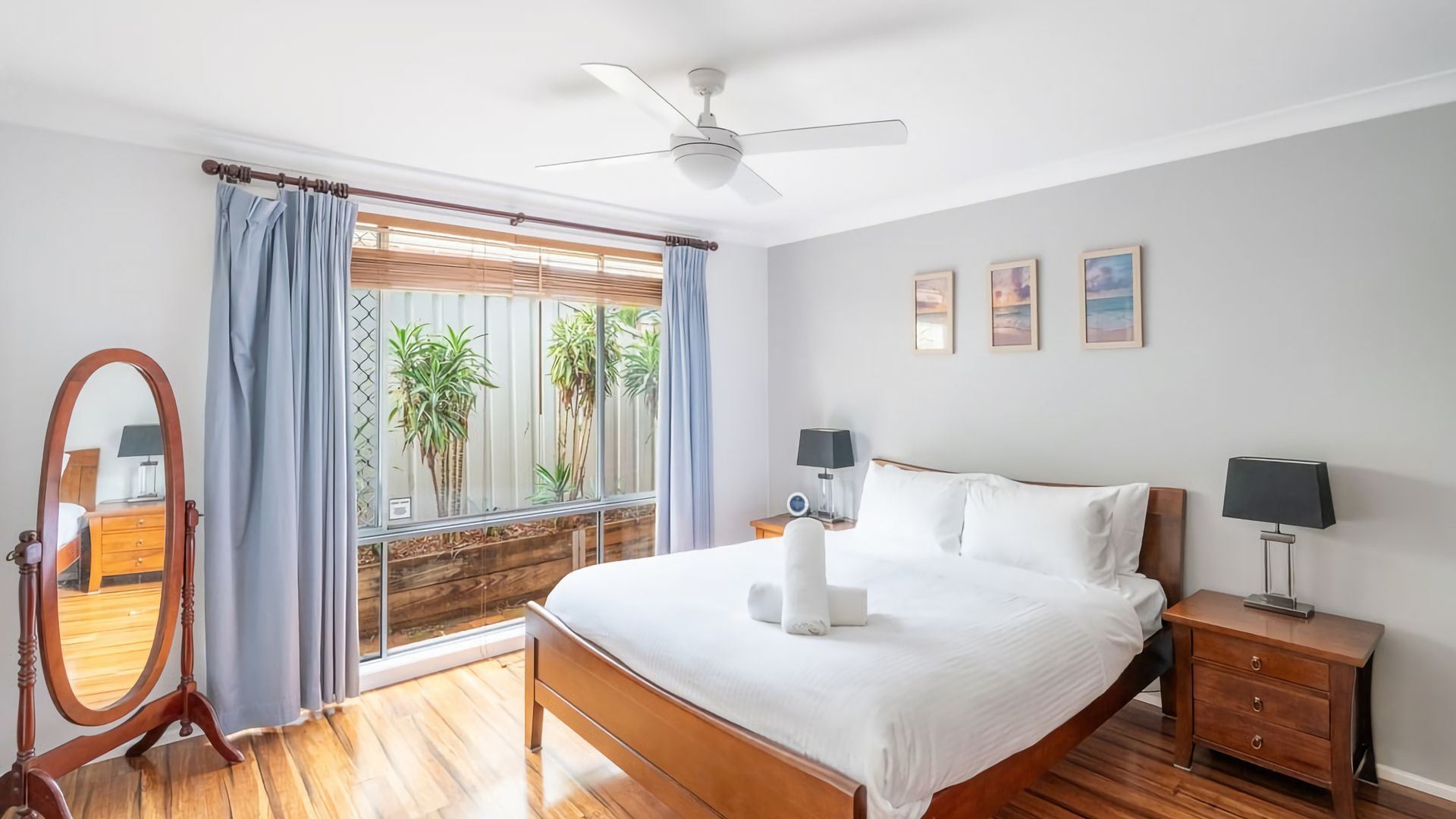 A bedroom with a large bed, wooden floor, ceiling fan, and a window with curtains.