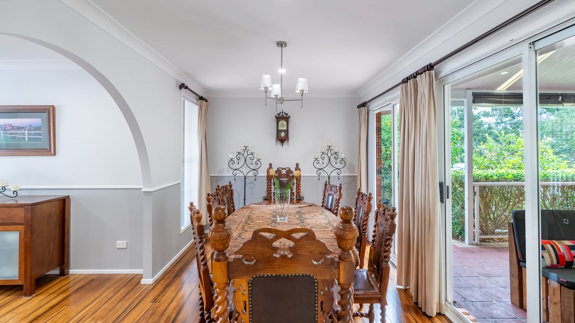 A dining area with a wooden table and chairs, a chandelier, and a view of greenery outside.