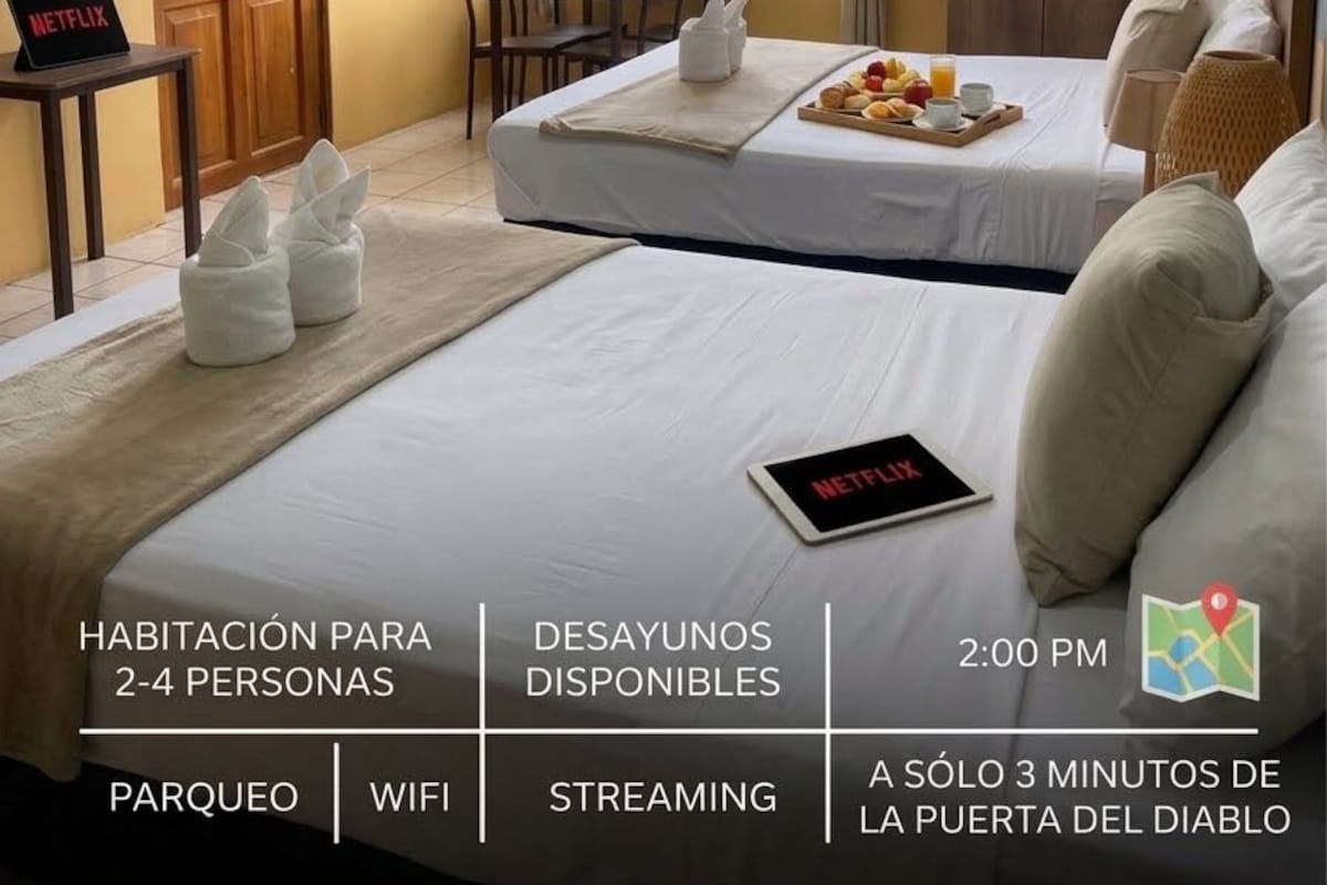Habitación de hotel con cama, escritorio, silla y bandeja de desayuno. Se observa un cartel que indica 'STREAMING DISPONIBLE' y otro que anuncia 'DESATANCO INCLUIDO'. La habitación tiene vistas al exterior.