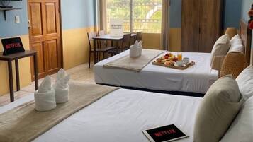 Habitación de hotel con cama, escritorio, silla y bandeja de desayuno. Se observa un cartel que indica 'STREAMING DISPONIBLE' y otro que anuncia 'DESATANCO INCLUIDO'. La habitación tiene vistas al exterior.