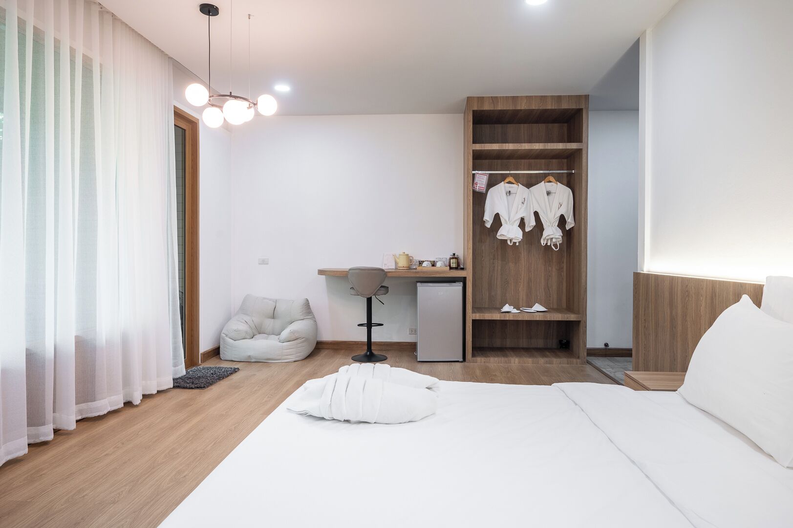 A modern hotel room with a bed, a desk with a computer, a chair, a wardrobe, and a hanging light fixture.