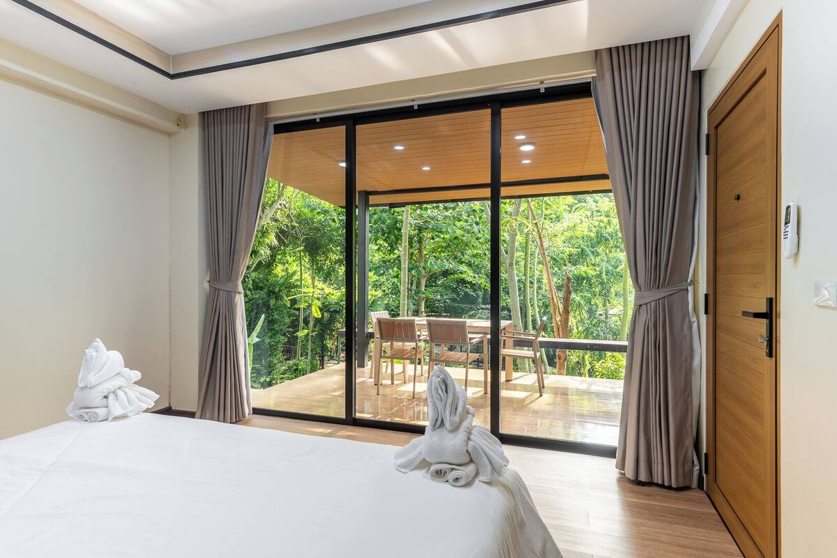 A bedroom with a large bed, a wooden door, and a sliding glass door leading to an outdoor seating area with a table and chairs.
