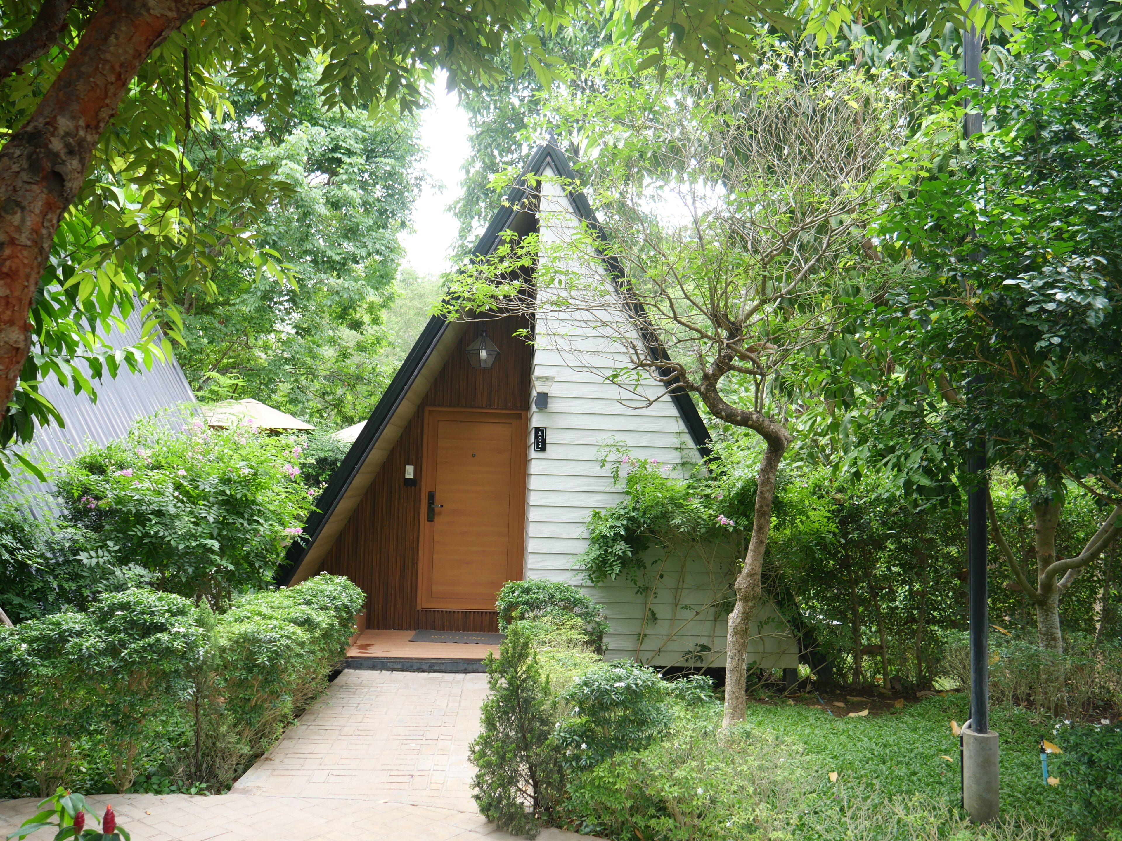 A small, wooden cabin with a sloped roof, surrounded by dense greenery and a paved pathway leading to the entrance.