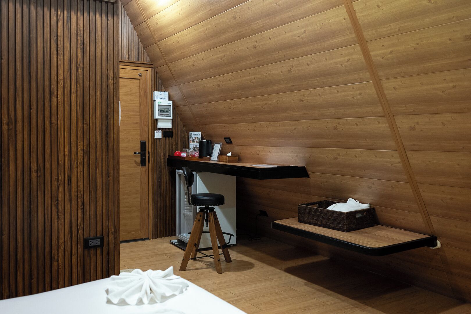 A wooden interior with a kitchenette, a stool, a small table, and a shelf with a box.