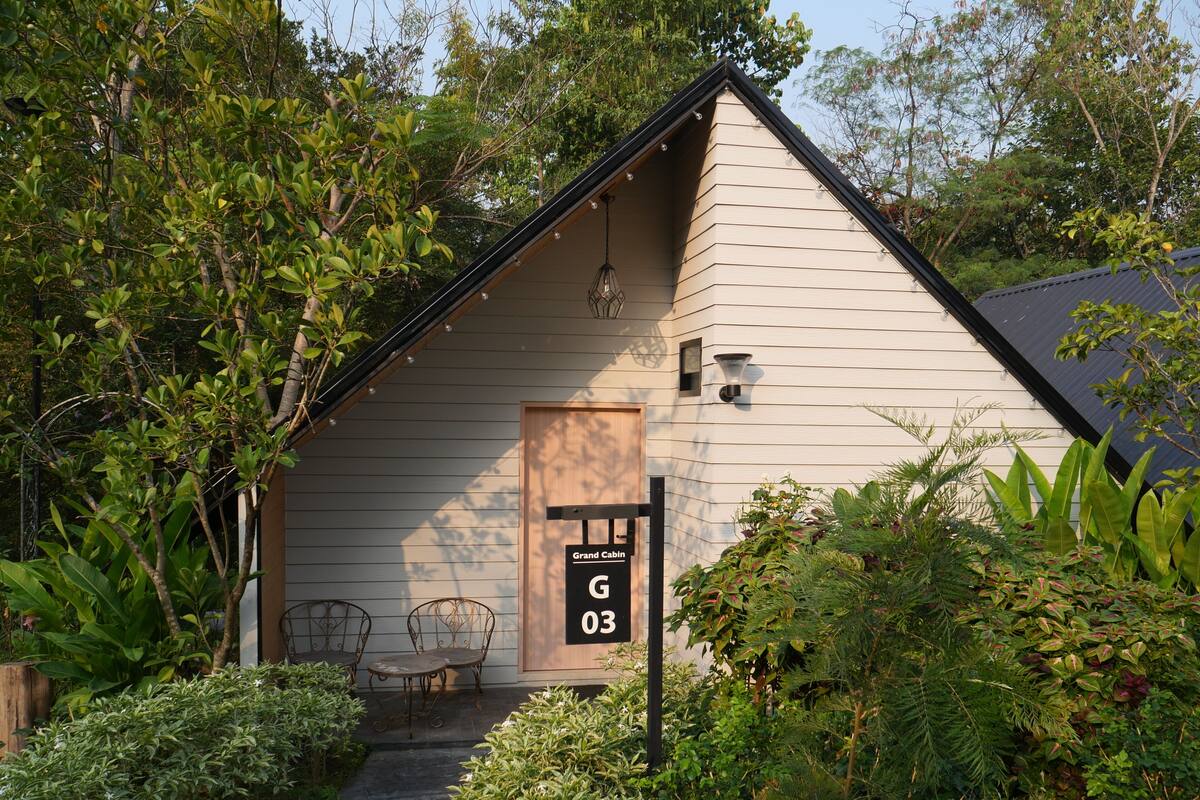 Una cabaña con puerta de madera, un cartel que indica 'Grand Cabin 03', y un pequeño espacio al aire libre con sillas de metal.