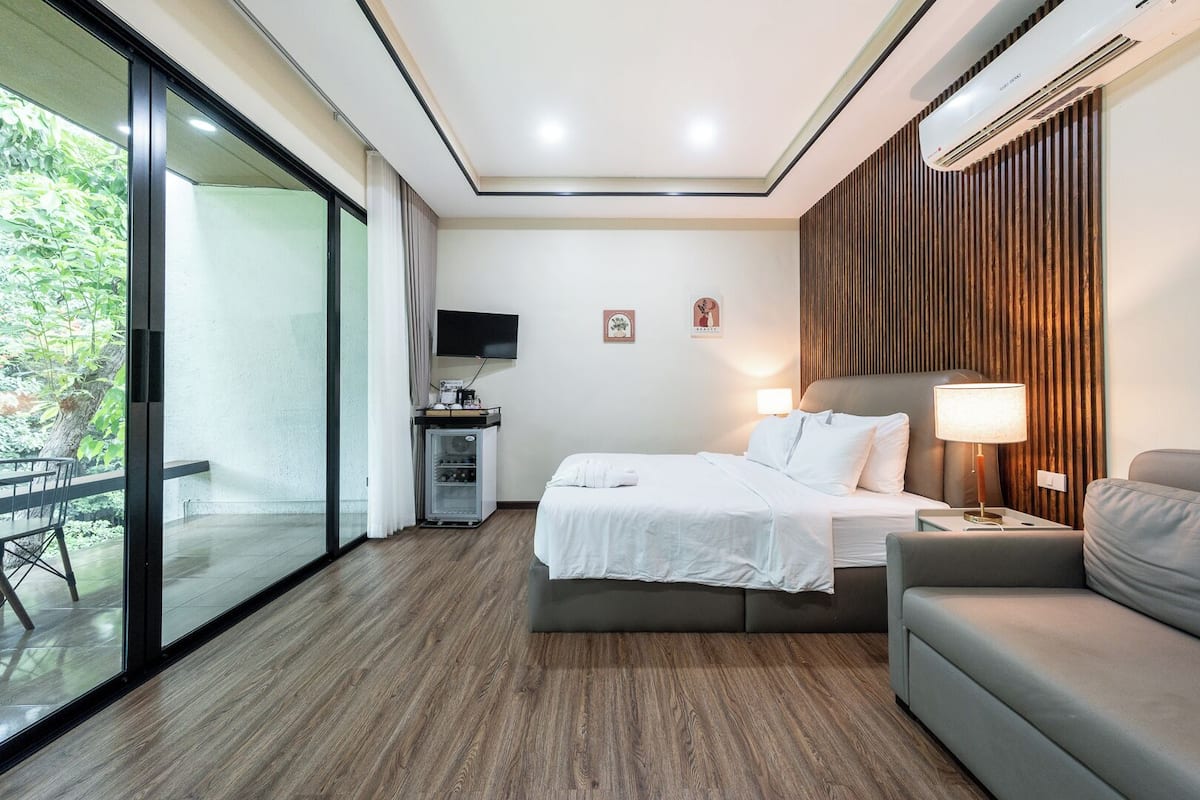 A hotel room with a bed, a sofa, a TV, a minibar, and a sliding glass door to a balcony.