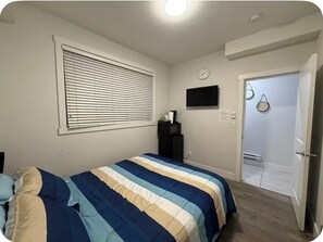 A bedroom with a bed, a window with blinds, a wall-mounted TV, a clock, and a door leading to another room. - Private room in surrey (Surrey)