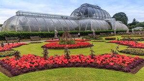 Un hivernacle gran amb estructura de cúpula, envoltat de llits de flors ben cuidats i camins.