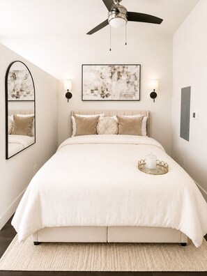 Une chambre à coucher comprenant un lit, un ventilateur de plafond, des lampes fixées au mur, un tableau abstrait encadré et un miroir.