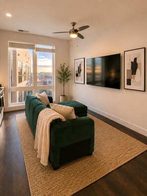 Un salon moderne avec un canapé, un ventilateur de plafond, une grande fenêtre et des œuvres d’art accrochées au mur.