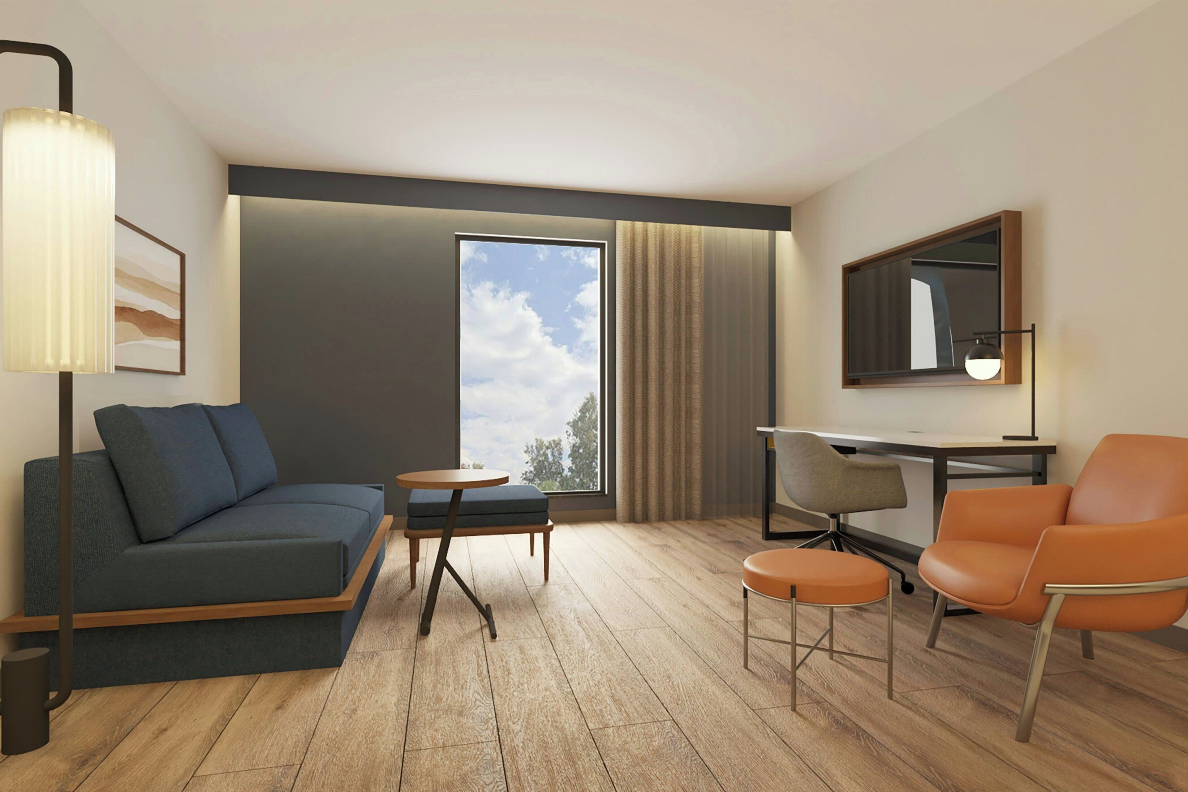 Una habitación de hotel moderna con sofá, una mesita redonda pequeña, una silla naranja, un escritorio y un gran ventanal con cortinas.