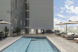 Un área de piscina moderna con sillones de descanso y sombrillas, rodeada por un edificio con amplios ventanales y plantas en macetas.