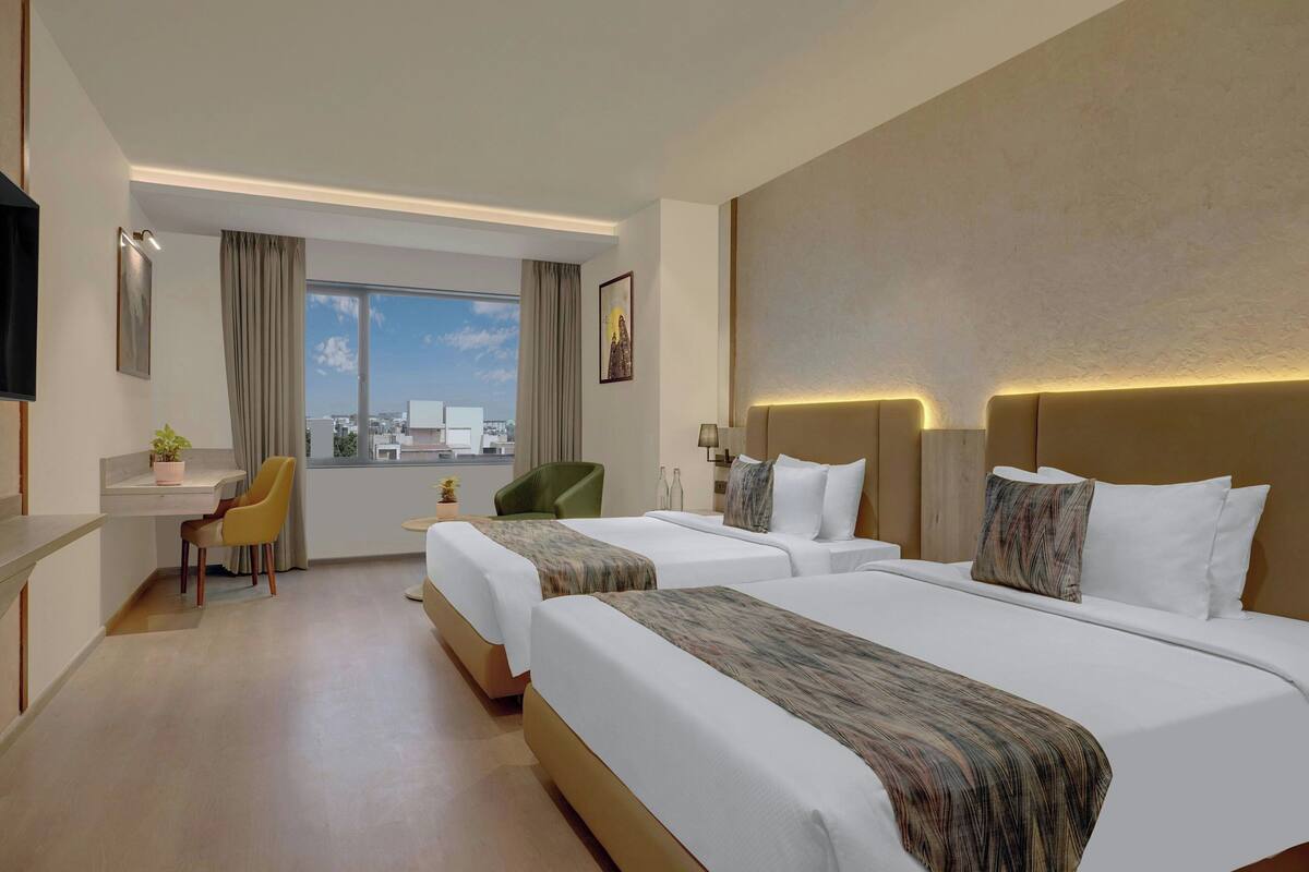 Quarto de hotel com duas camas, uma escrivaninha, uma cadeira, televisão e janela com vista para a cidade.