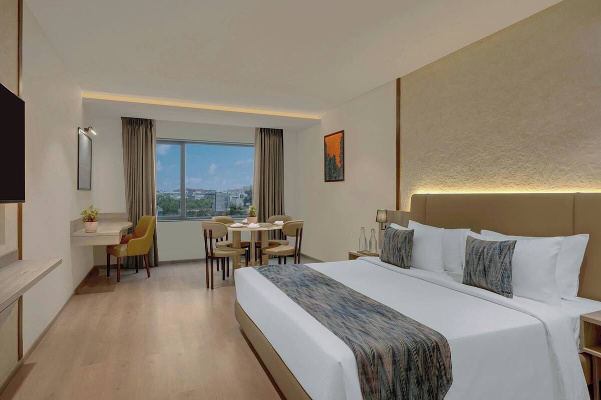 Quarto de hotel com uma cama grande, uma escrivaninha com cadeira, uma mesinha e vista para a cidade.