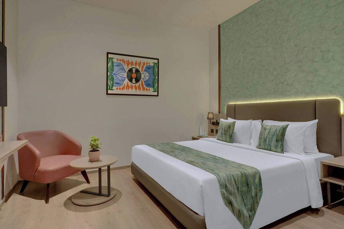 Quarto de hotel com uma cama, uma cadeira, uma mesinha e uma obra de arte na parede.