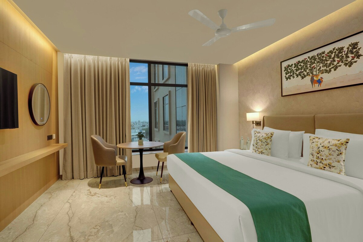 A hotel room with a bed, a desk, two chairs, a ceiling fan, a large window with curtains, and a wall-mounted TV.