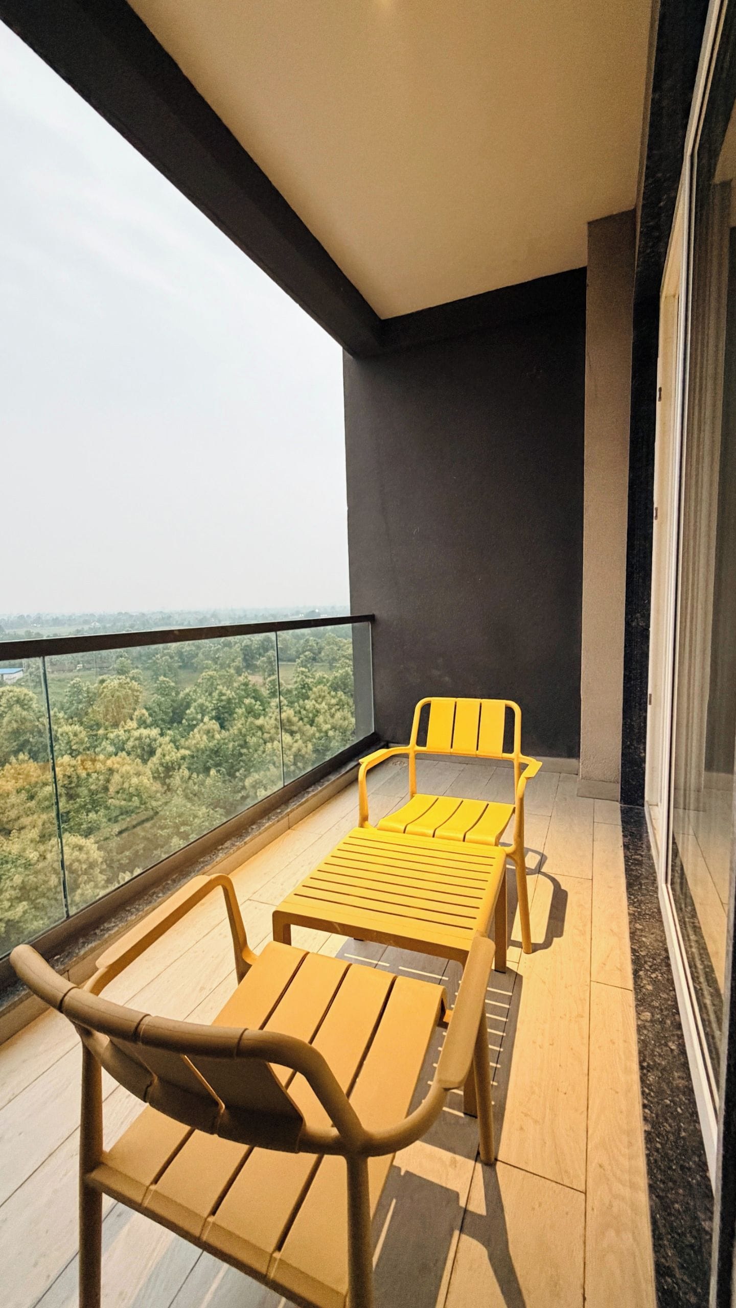 A balcony with a table and two chairs, a view of greenery, and a dark wall.