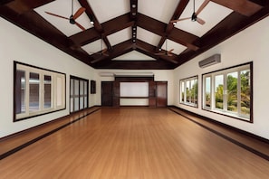 Una habitación amplia con suelo de madera, ventanas grandes, ventiladores de techo y un equipo de aire acondicionado.